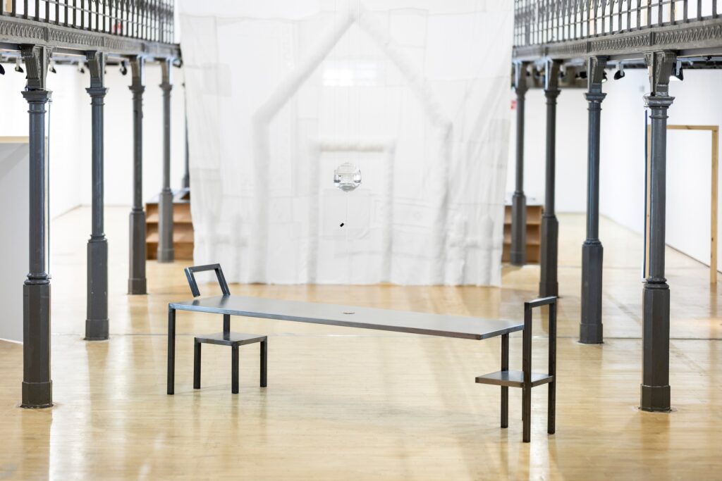 Architecture of Care, vue d’installation au Botanique : table en acier, chaises soudées, ampoule de verre suspendue