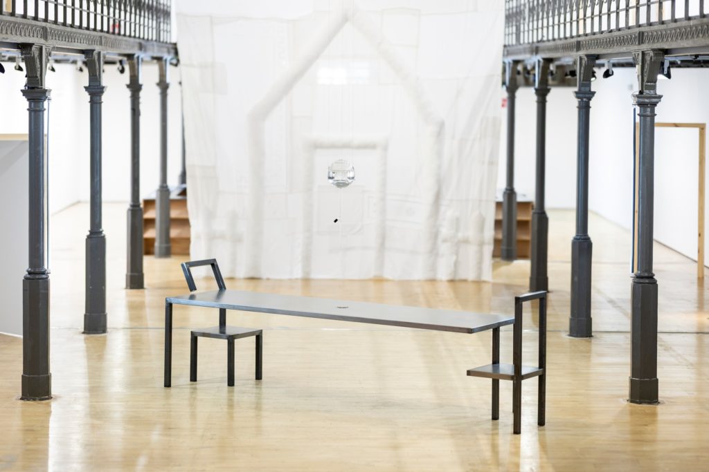 Architecture of Care, vue d’installation au Botanique : table en acier, chaises soudées, ampoule de verre suspendue